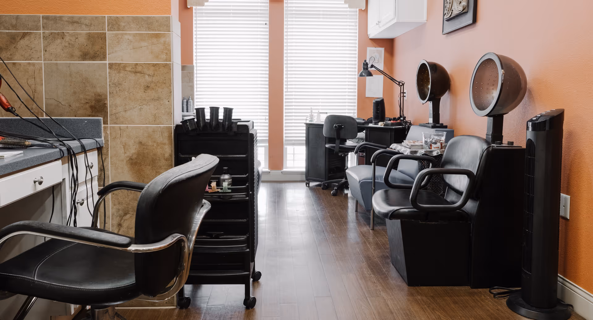 Interior view of a hair salon area with black salon chairs, hair dryers, a rolling cart with hair products, and a workstation with a lamp and supplies. The walls are painted orange and there is a window with blinds in the background.