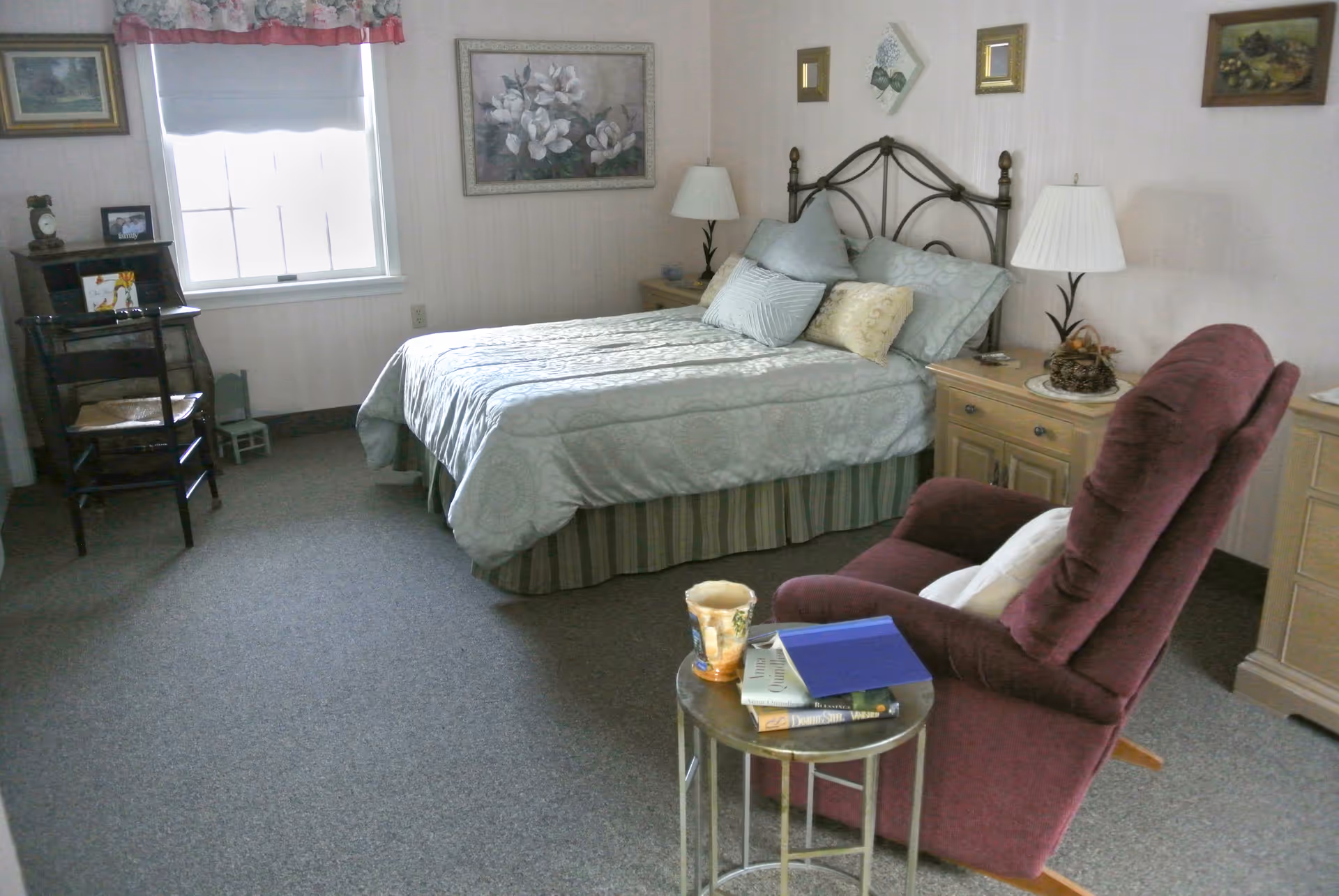 A cozy bedroom with a neatly made bed featuring multiple pillows, two bedside tables with lamps, a burgundy recliner chair next to a small round table holding books and a cup, a window with a floral valance, and framed artwork on the walls.