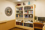 Small interior reading nook with a large bookshelf filled with books, a desk with a computer and chair, and a decorative round mirror on the wall.
