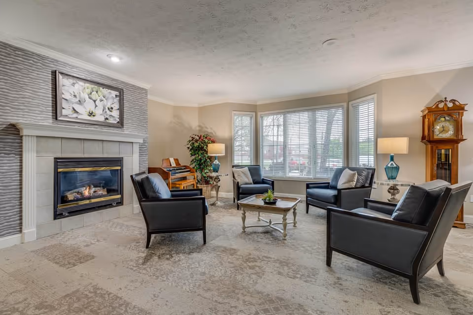 Bright sitting room with a fireplace, four leather armchairs around a coffee table, bay windows, a piano, and a grandfather clock.