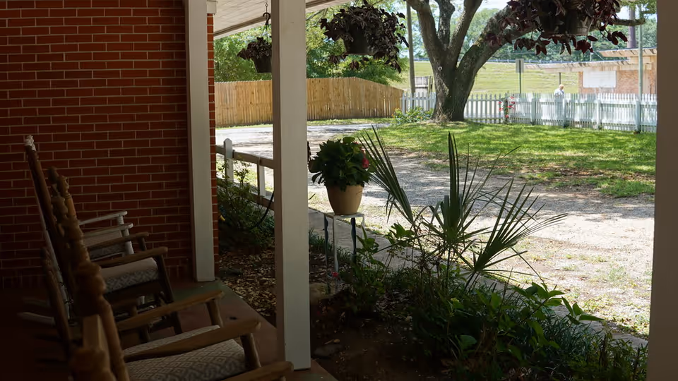 View from a covered porch with wooden rocking chairs looking out onto a garden area with plants, a tree, a gravel path, and a white picket fence in the background.