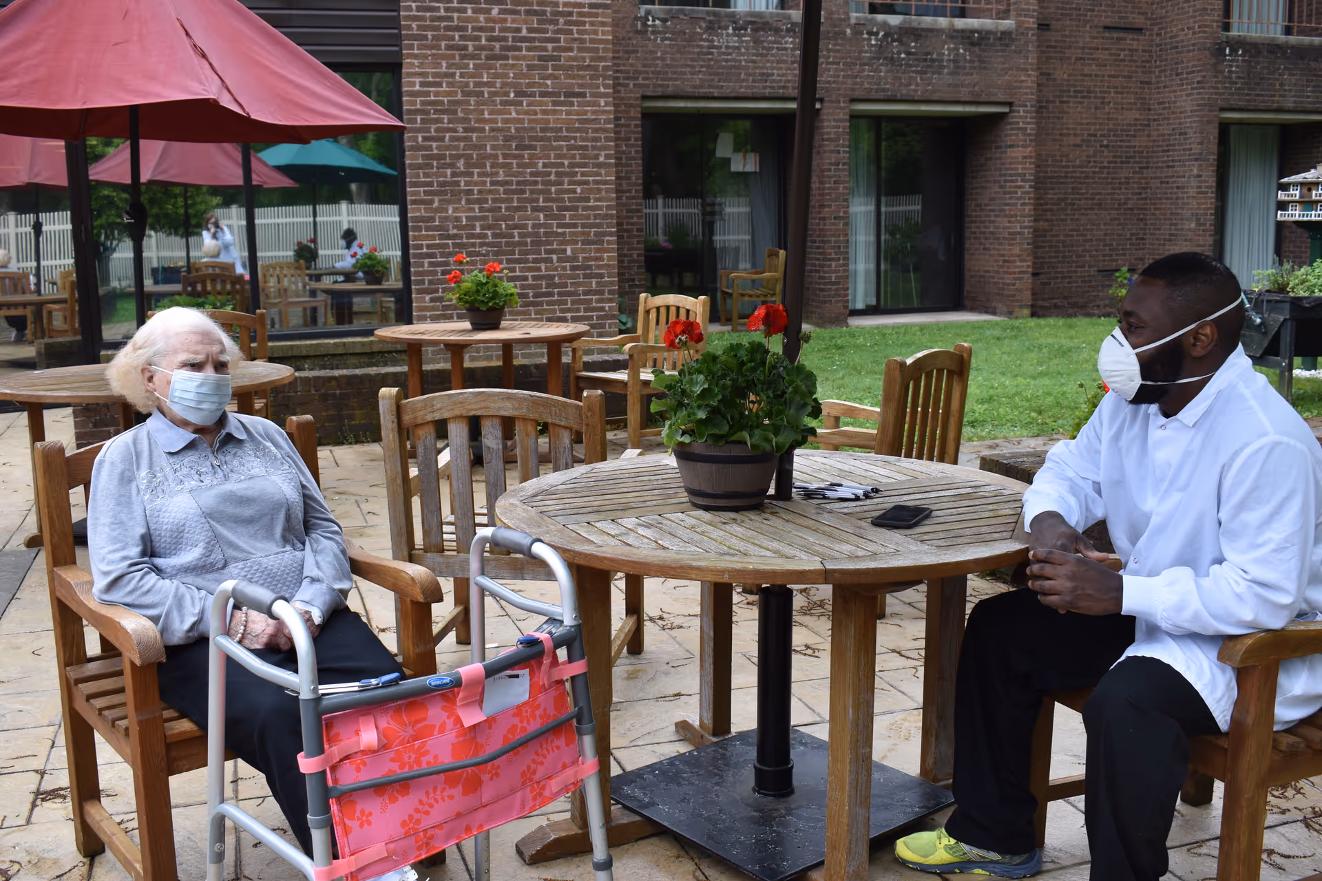 An elderly woman wearing a face mask sits on a wooden chair outdoors with a walker in front of her. Across from her, a man wearing a white shirt, black pants, yellow shoes, and a face mask sits on another wooden chair. They are seated at a round wooden table with a potted plant in the center. The setting is a patio area with additional tables, chairs, and umbrellas, adjacent to a brick building.