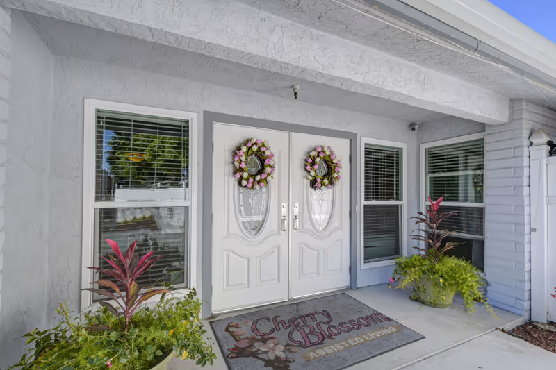 Entrance to Cherry Blossom Assisted Living featuring double white doors with glass panels, each decorated with a floral wreath. Two windows with blinds flank the doors. Potted plants are placed on either side of the entrance, and a welcome mat with the facility name and floral design lies in front of the doors.