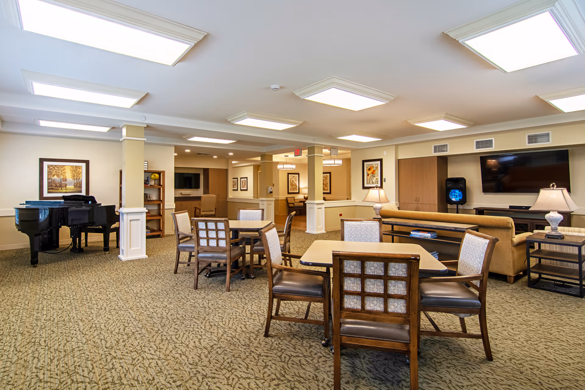 A spacious senior living common area with multiple tables and chairs, a grand piano in the corner, a large flat-screen TV mounted on the wall, and a comfortable sofa with side tables and lamps. The room has carpeted flooring, framed artwork on the walls, and bright ceiling lights.