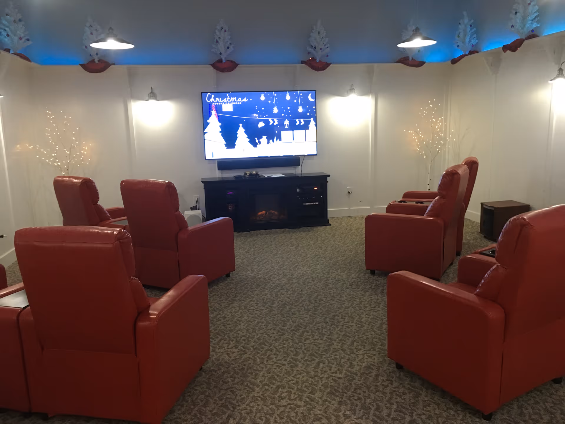 A cozy common room with six red recliner chairs arranged in two rows facing a wall-mounted flat screen TV displaying a Christmas-themed screen. The room has soft lighting with wall sconces and decorative white trees with lights on either side of the TV. The ceiling has blue accent lighting and white holiday decorations along the top edge of the walls. The floor is carpeted with a patterned design.