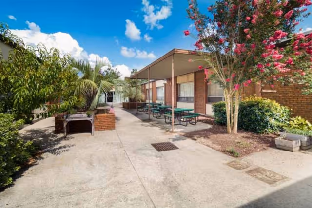 Outdoor patio area at Azalea Health Center with concrete walkway, green picnic tables under a covered area, surrounded by trees and bushes including a flowering tree with pink blossoms, under a blue sky with some clouds.