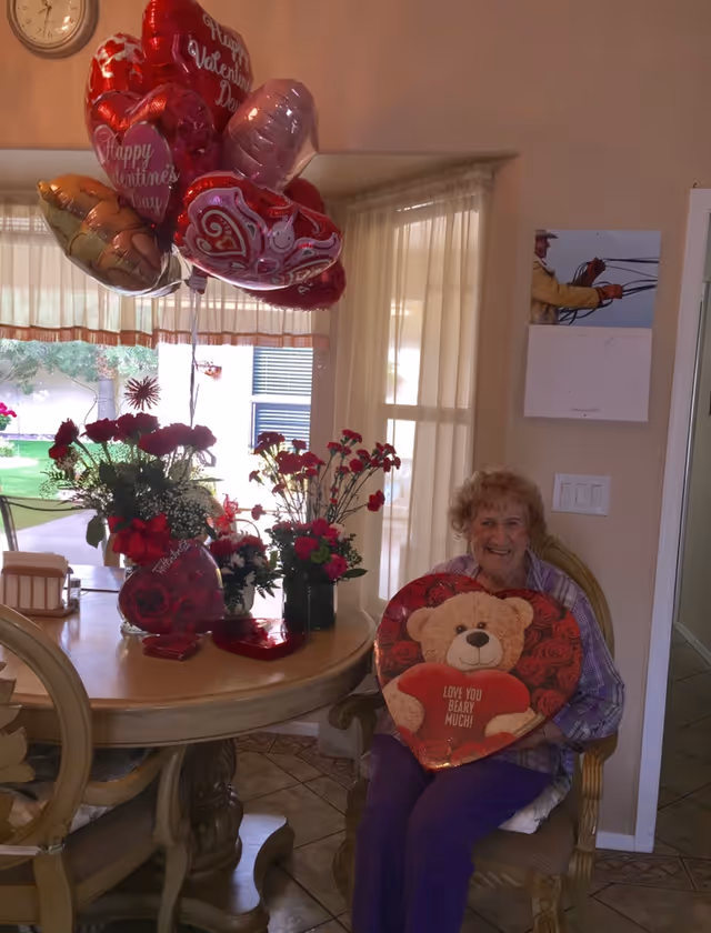 An elderly woman sitting on a chair in a dining area holding a heart-shaped Valentine’s Day balloon with a teddy bear and the message 'Love you beary much!'. On the round dining table next to her are several bouquets of red flowers and multiple Valentine’s Day themed balloons. A window with sheer curtains and a calendar on the wall are visible in the background.
