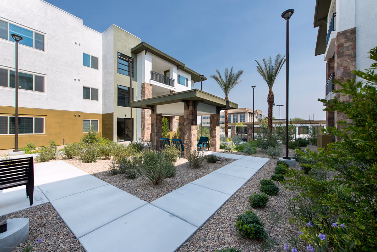 Sunlit landscaped courtyard with walkways, seating and a covered patio between multi-story residential buildings and palm trees.