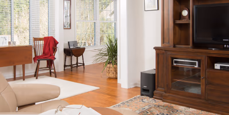 Sunlit living room with hardwood floors, a large wooden entertainment cabinet, a leather armchair, and a chair with a red throw by the windows.