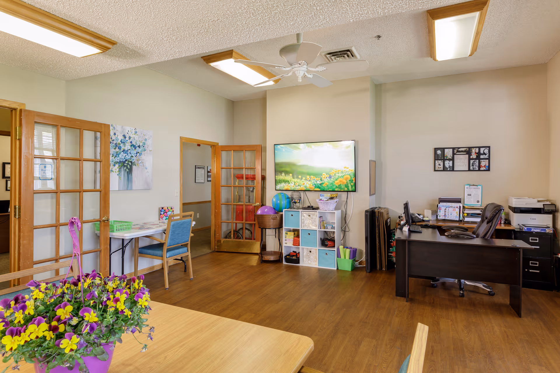 A well-lit office and common area in an assisted living facility featuring a wooden desk with office supplies and a computer, a wall-mounted TV displaying a scenic image, a small storage unit with colorful bins, a table with chairs, and a purple flower pot with yellow and purple flowers in the foreground. The room has wooden flooring, beige walls, and glass-paneled wooden doors.