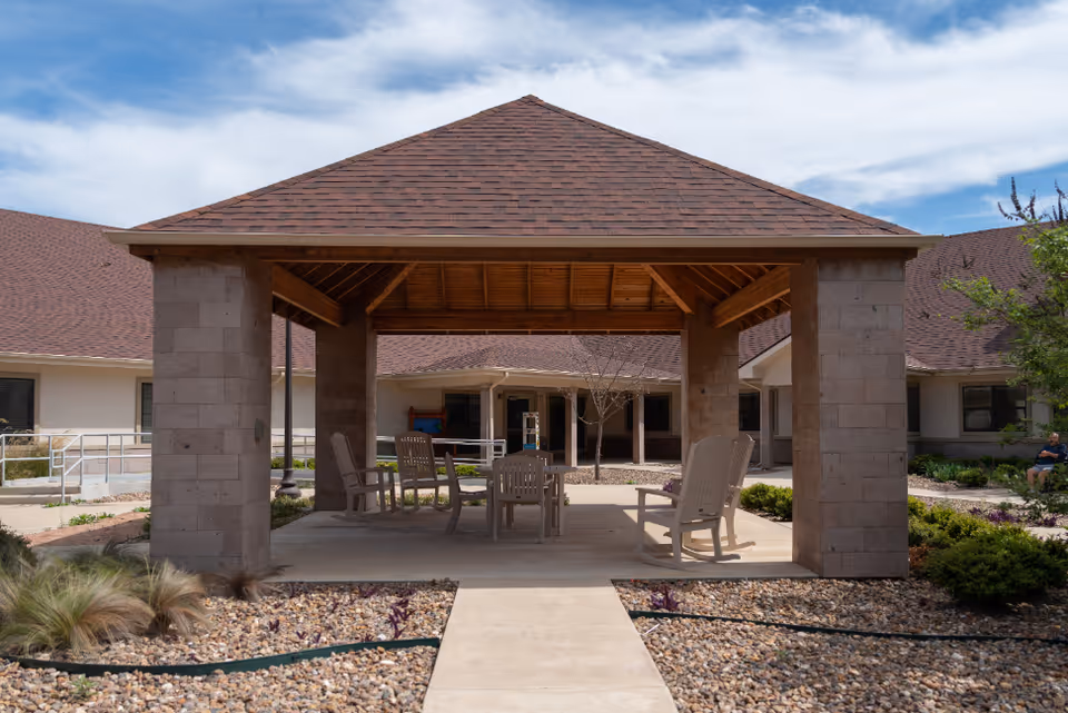 Outdoor covered seating area with a wooden roof supported by stone pillars, surrounded by a landscaped garden with gravel and plants. Several chairs and a table are arranged under the shelter. The background shows a single-story building with a brown shingled roof and windows.