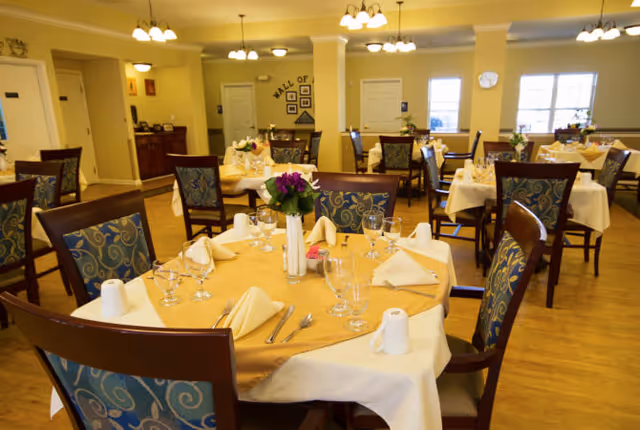 A dining room with multiple round tables covered with yellow tablecloths and white overlays, each set with folded napkins, glassware, and silverware. The chairs have wooden frames with blue patterned cushions. The room is well-lit with ceiling lights and has large windows letting in natural light. There are decorative flowers on the tables and a wall in the background with framed pictures and a sign that reads 'Wall of'.