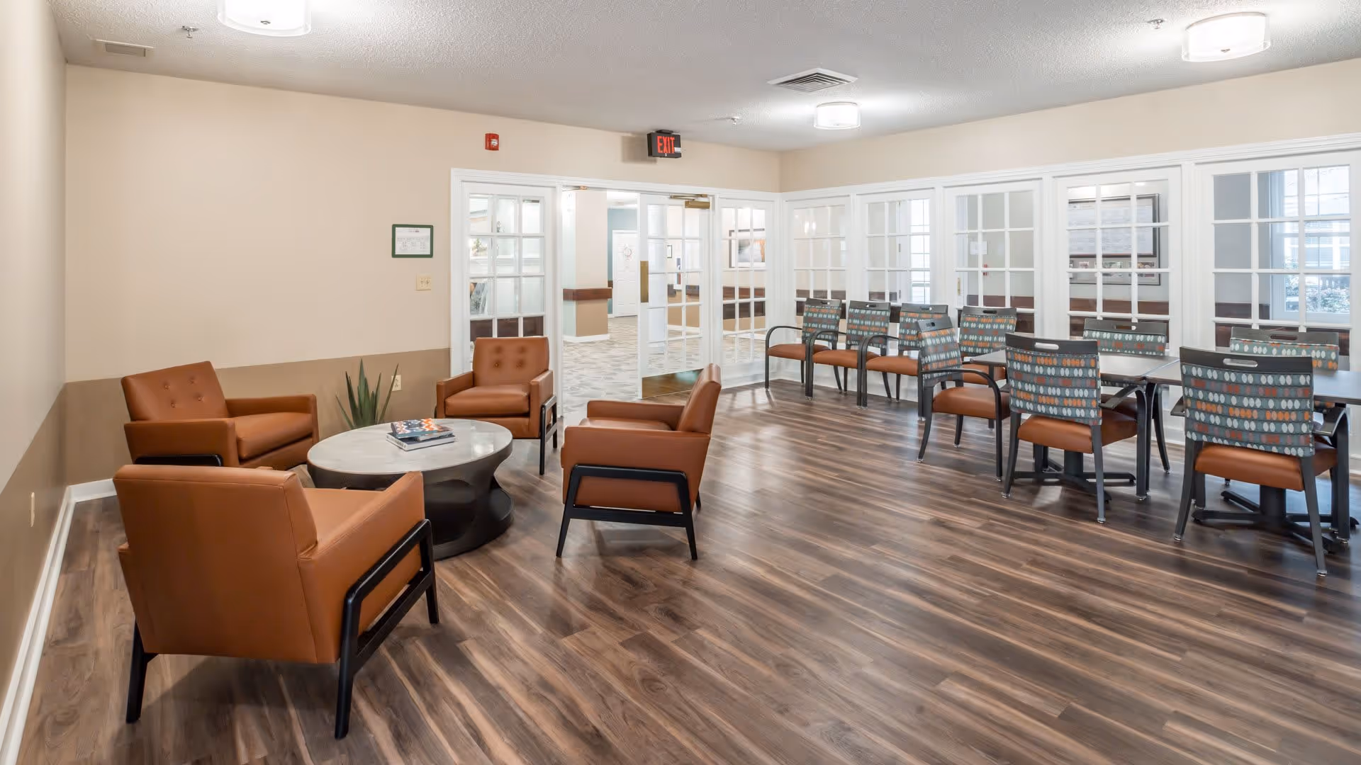 A spacious common area with wood flooring featuring a seating arrangement of four brown armchairs around a round coffee table on the left side, and several tables with patterned chairs on the right side. The room has beige walls with white trim and large windows with white frames, allowing natural light to enter. There are ceiling lights and an exit sign above a set of double glass doors in the background.