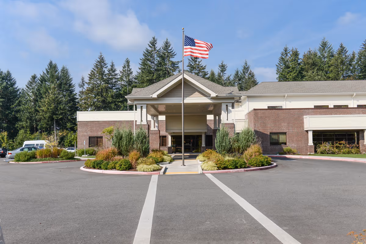 Front exterior view of Lacey Post Acute & Rehabilitation building with an American flag on a flagpole in the center, surrounded by landscaped bushes and trees, under a partly cloudy blue sky.