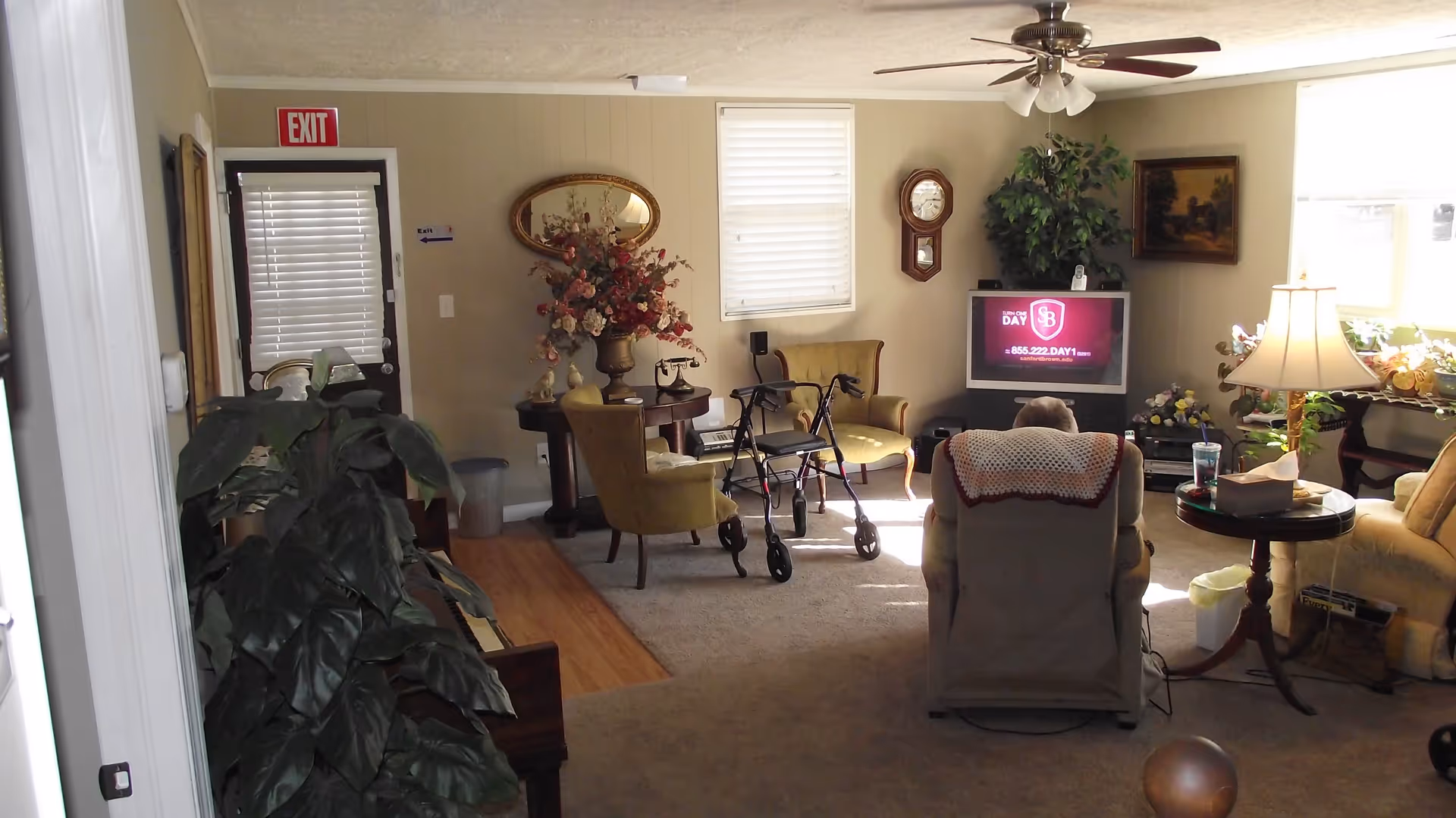 A cozy living room with beige walls and carpet. There is a person sitting in a recliner chair facing a TV, which displays an advertisement. The room contains several chairs, a walker, a round table with a floral arrangement, a floor lamp, a wall clock, and various plants. A ceiling fan with lights is mounted on the ceiling, and there is an exit door with a red exit sign above it.