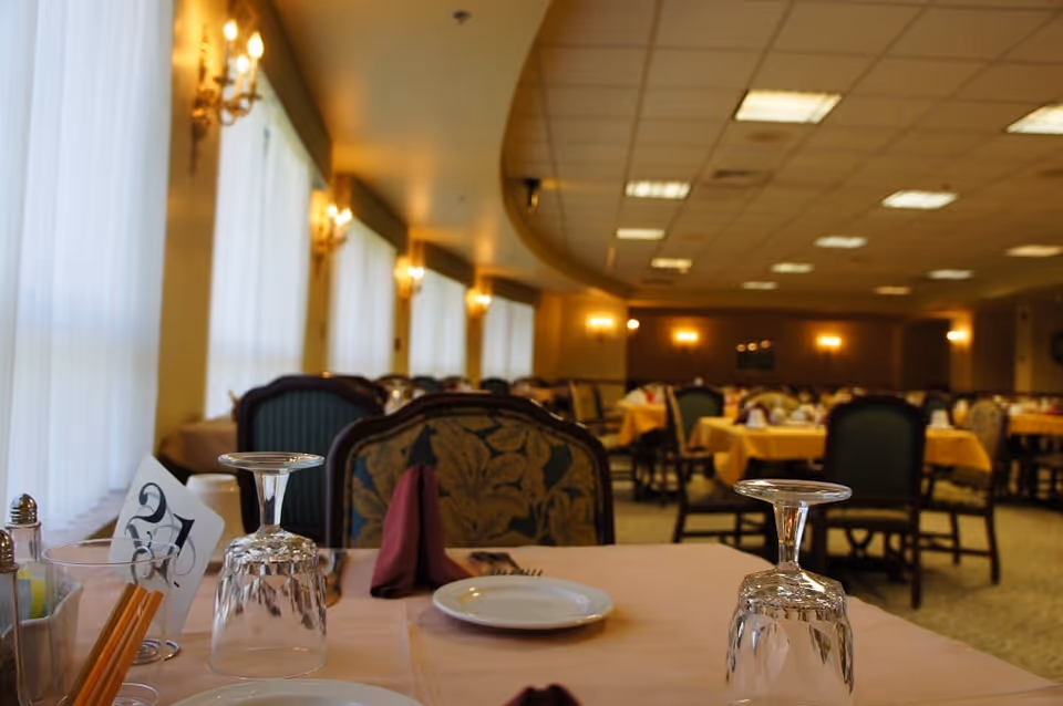 An elegantly set dining table with inverted wine glasses and plates in a large, softly lit dining room with many tables and chairs.