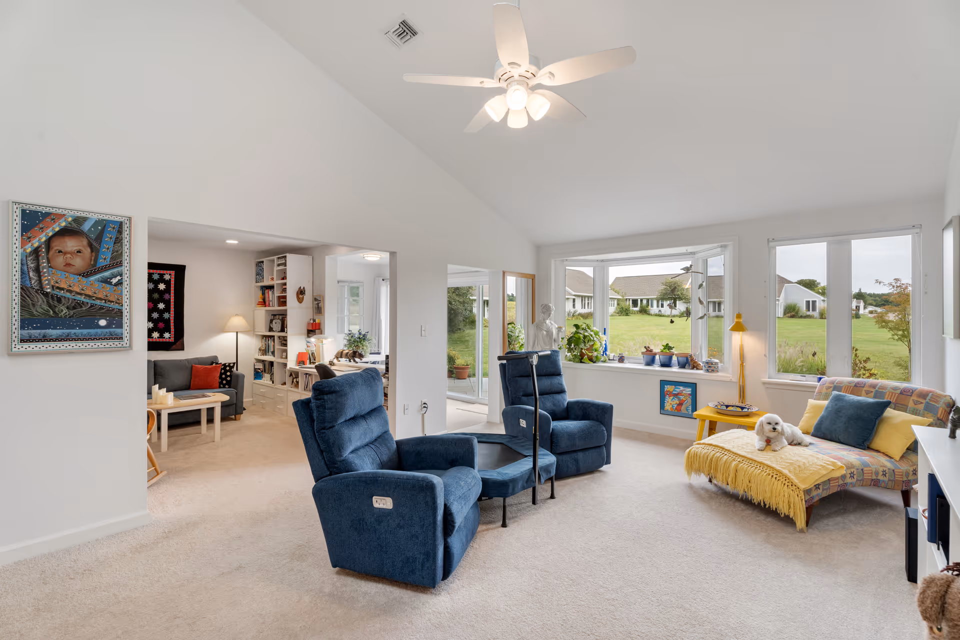 A bright living room with two blue recliner chairs and a colorful chaise lounge with a small white dog resting on it. The room has large windows showing a green outdoor view, a yellow side table with a lamp, and various plants on the windowsill. In the background, there is an adjacent room with a gray sofa, a wooden coffee table, bookshelves, and framed artwork on the walls.