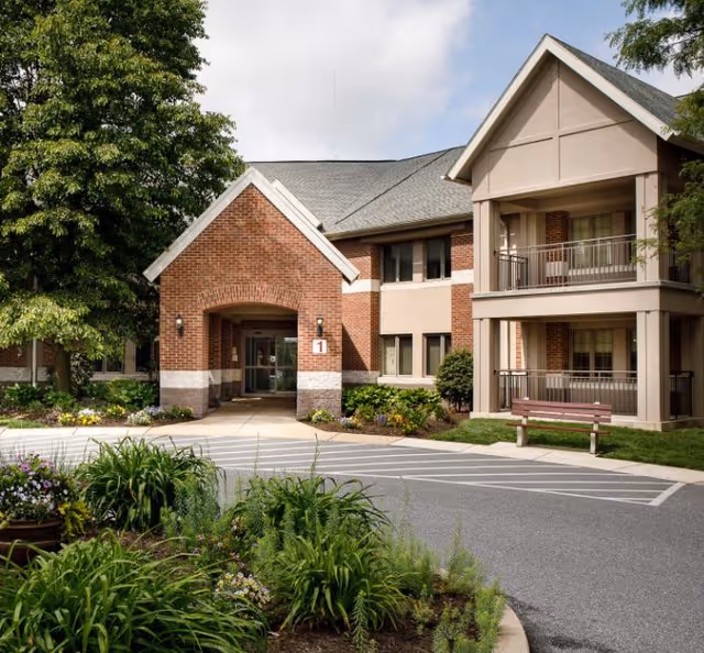 Exterior view of a senior living facility building with a brick and beige facade, featuring an entrance with a covered walkway, balconies, a bench, and landscaped greenery including trees and flowering plants.