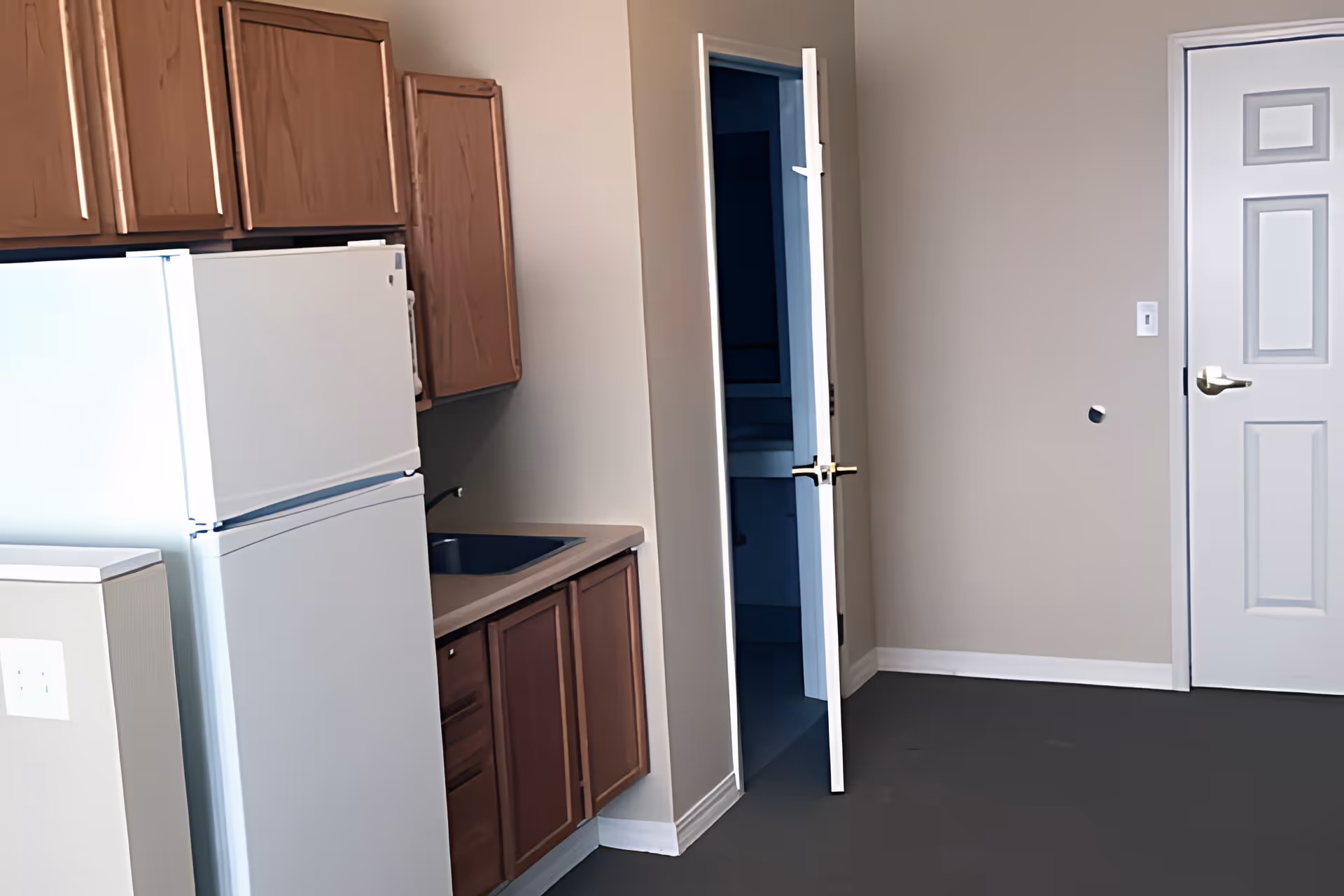 Interior view of a small kitchen area with wooden cabinets, a white refrigerator, and a countertop with a sink. There is an open door leading to another room and a closed white door on the right side.