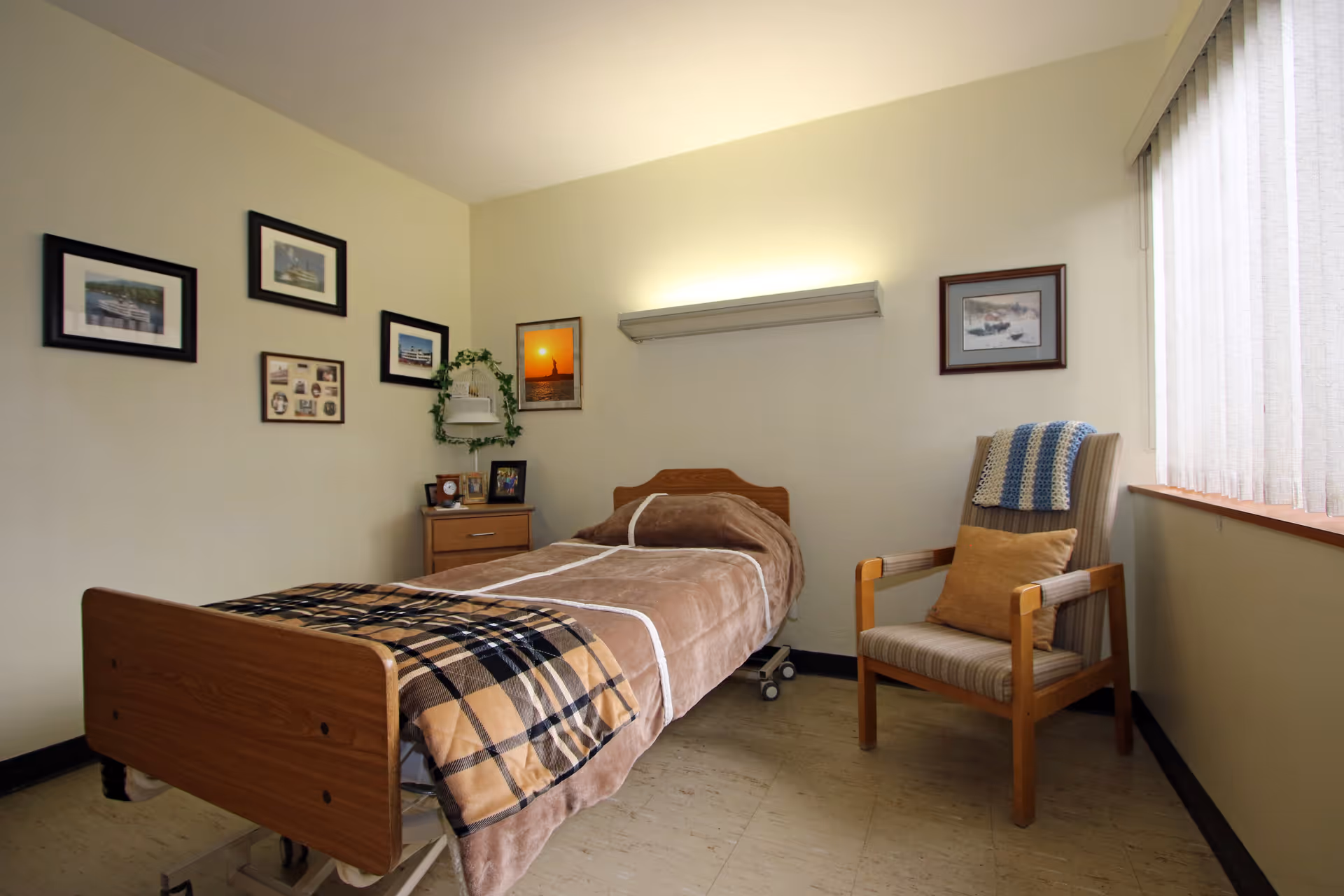 A simple bedroom with a single hospital-style bed covered with a brown blanket and a plaid throw. There is a wooden nightstand with framed photos and a plant on it. Several framed pictures hang on the walls. A wooden armchair with a cushion and a crocheted blanket is placed near a window with vertical blinds.