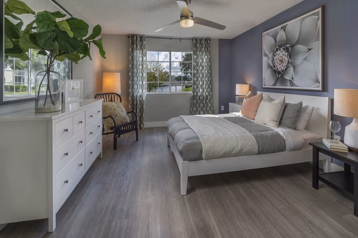 A modern bedroom with a white bed frame and gray and white bedding, decorated with multiple pillows. There is a large framed succulent artwork on the wall above the bed. On the right side of the bed is a black nightstand with a white lamp, a glass of water, and some books. On the left side of the room, there is a white dresser with multiple drawers, a large glass vase with green leafy branches, and a black chair with a fluffy cushion. The room has a large window with patterned curtains, gray walls, and wood flooring. A ceiling fan with a light is mounted on the ceiling.