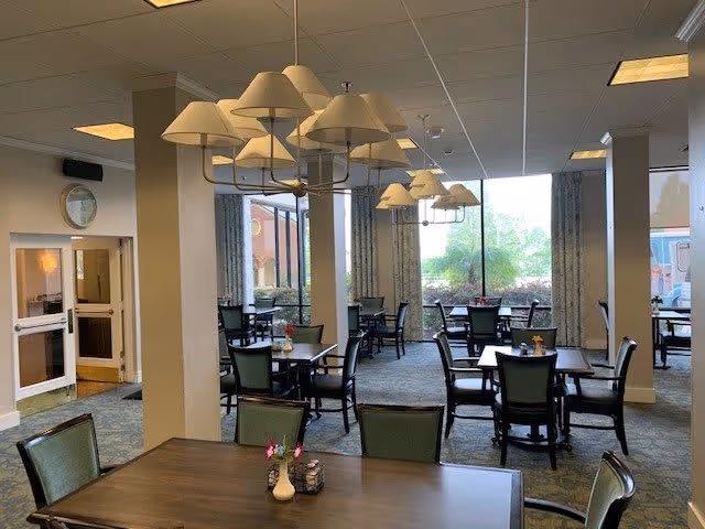 Interior view of a dining room in a senior living facility with multiple tables and chairs arranged neatly. The room has large windows letting in natural light, patterned curtains, and ceiling lights with multiple lampshades. Small flower vases are placed on the tables.