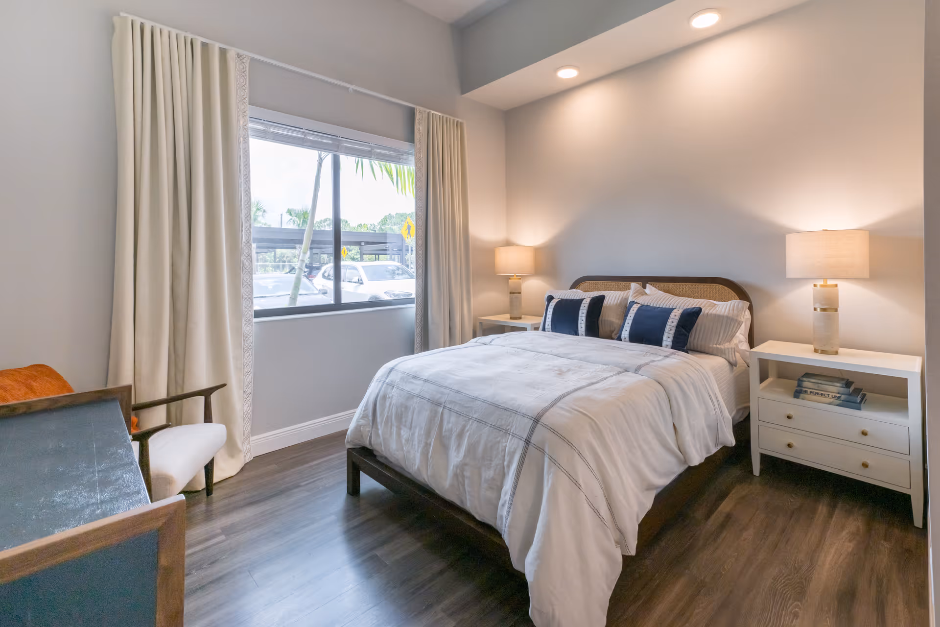 A neatly made bed with white and blue pillows in a bright bedroom featuring two bedside tables with lamps, a window with cream curtains, and a wooden floor.
