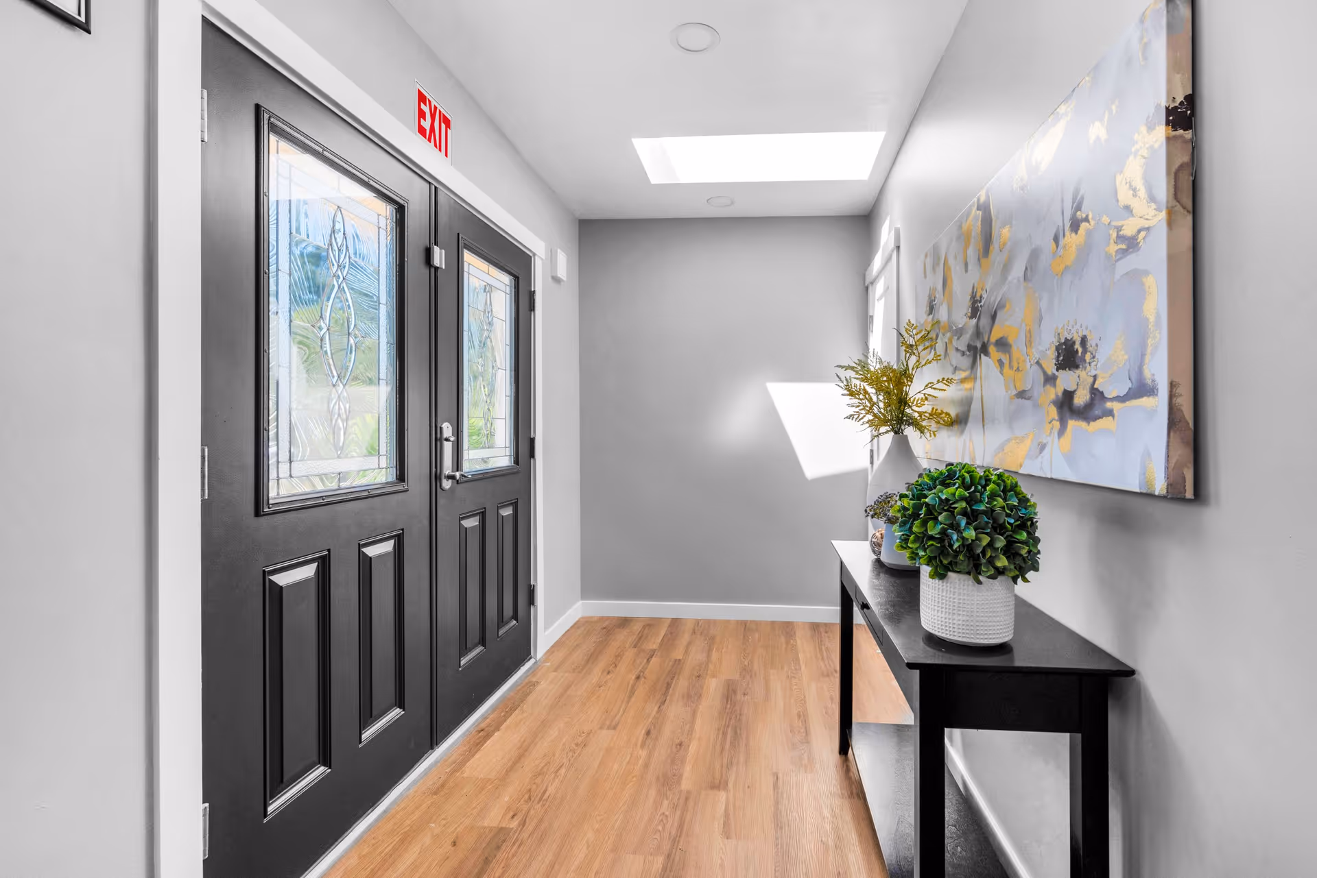 A bright entry hallway with black double doors featuring decorative glass, a console table topped with potted plants, and large wall artwork.