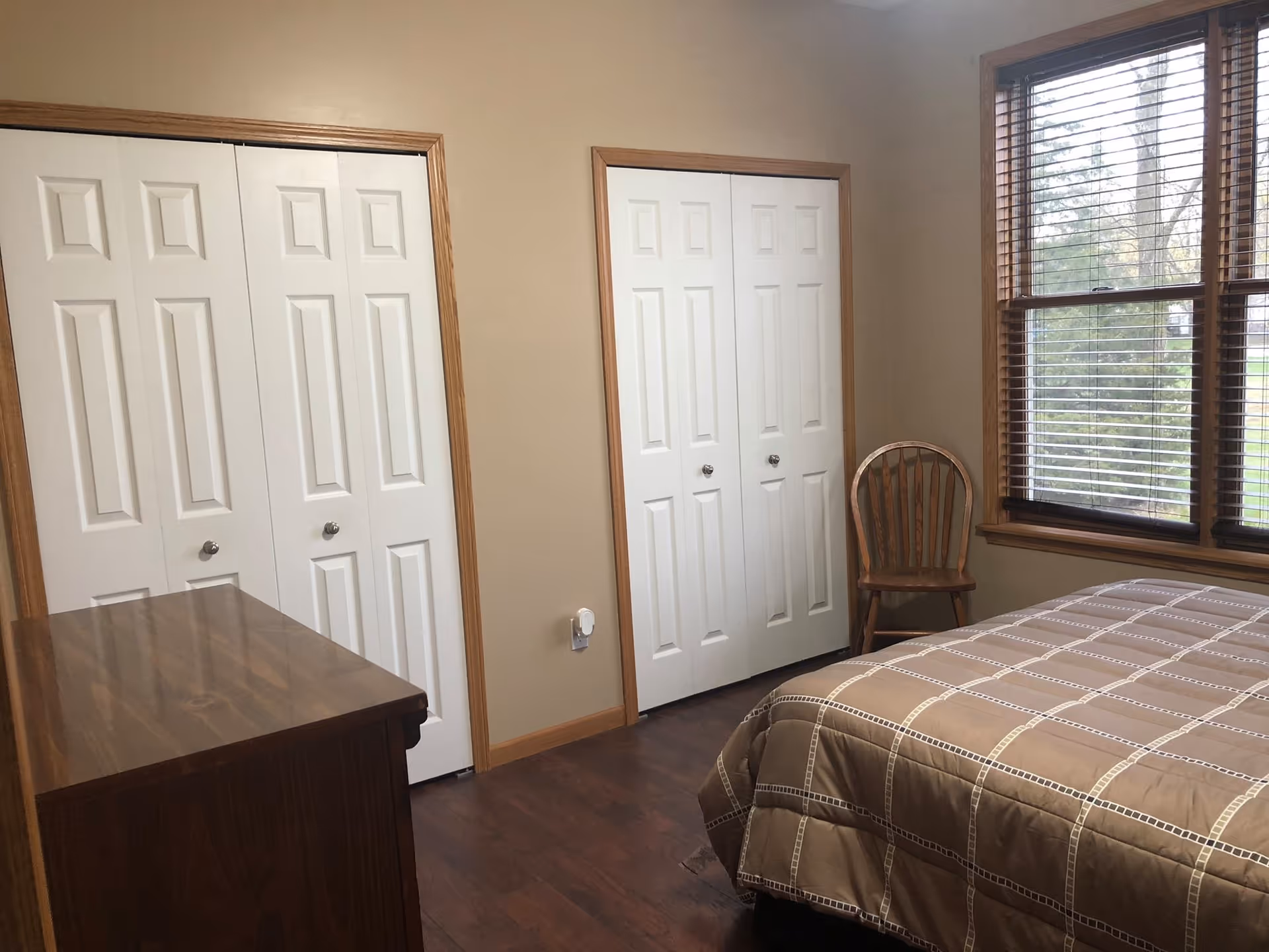 A bedroom with a bed covered in a beige checkered comforter, a wooden chair near a window with wooden blinds, two white double-door closets with wooden trim, and a wooden dresser. The walls are painted beige and the floor is dark wood.