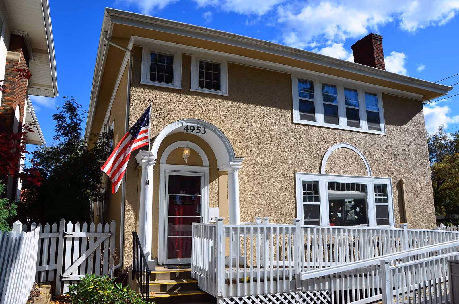 Exterior view of a two-story beige stucco building with white trim, featuring a red front door under an arched entryway with the number 4953 above it. An American flag is mounted near the entrance, and a white picket fence with a gate and a wheelchair ramp is visible in front of the building. The sky is blue with some clouds.