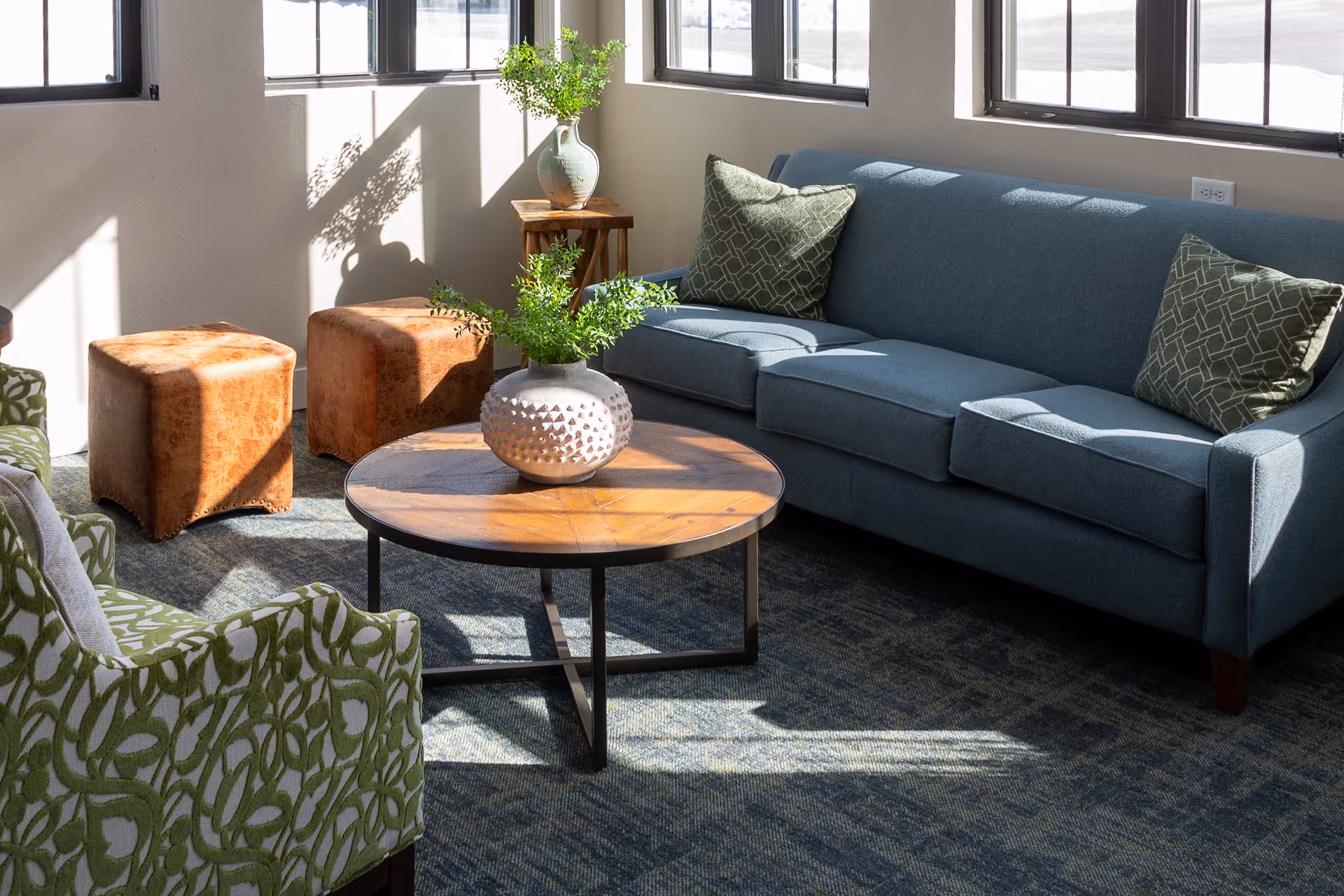 A bright living room area with a blue sofa adorned with two patterned green pillows, two brown leather ottomans, a round wooden coffee table with a textured white vase holding green foliage, and a green patterned armchair. Sunlight streams through large windows casting shadows on the carpeted floor.