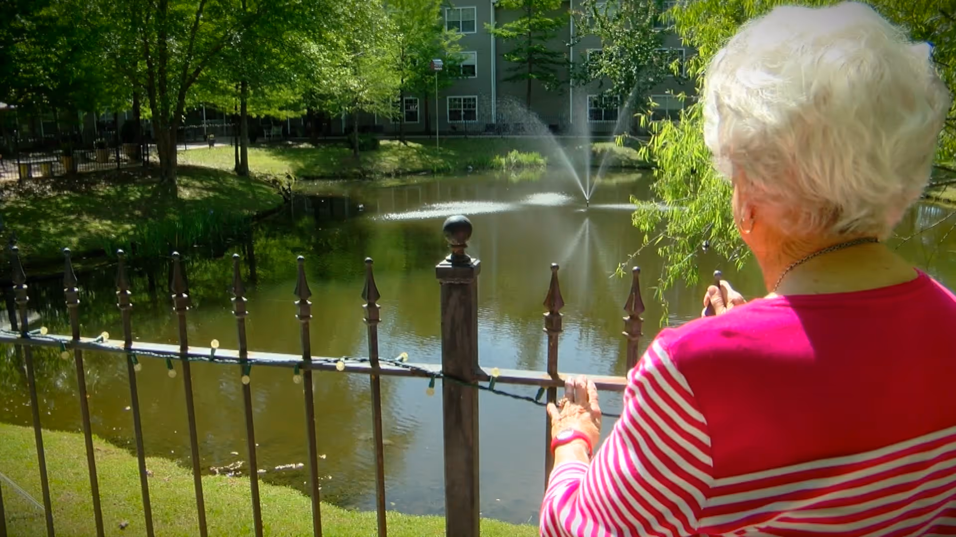 Person with white hair wearing a pink striped top stands by an iron fence overlooking a pond with a fountain and trees.