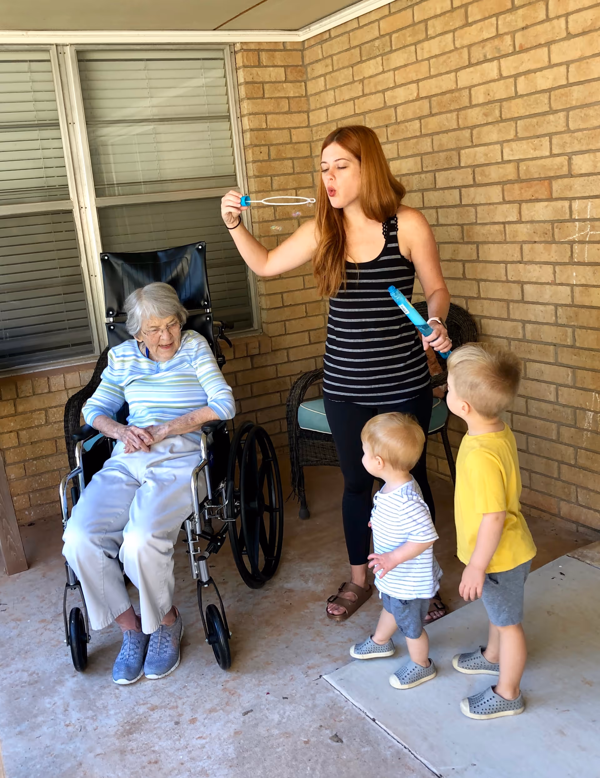 An elderly woman in a wheelchair is sitting outside on a covered patio with brick walls. A young woman is blowing bubbles while two small children watch the bubbles with interest.