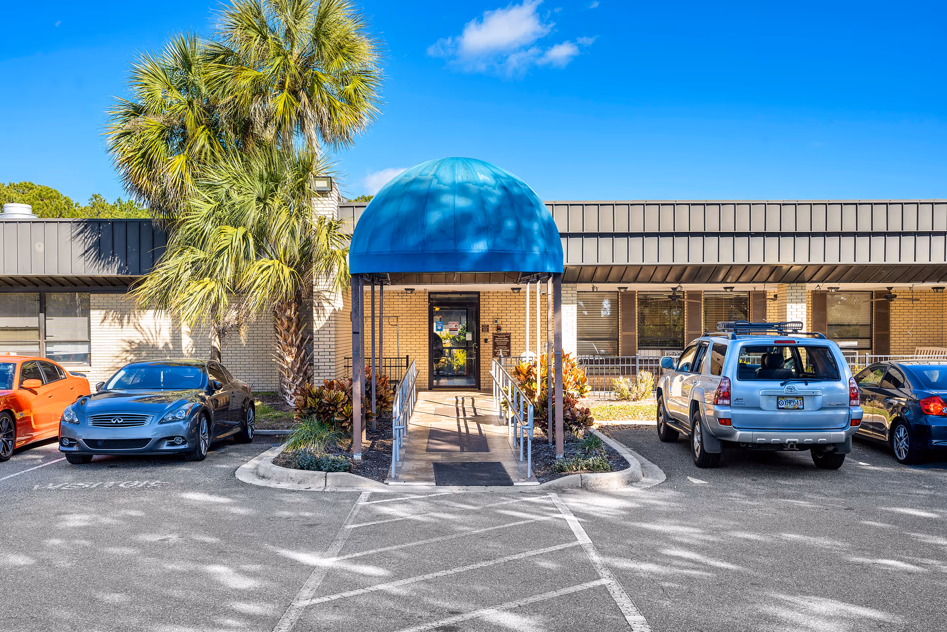 Entrance to Fernandina Beach Rehab and Nursing Center with a blue canopy over a wheelchair-accessible ramp, surrounded by parked cars and palm trees under a clear blue sky.