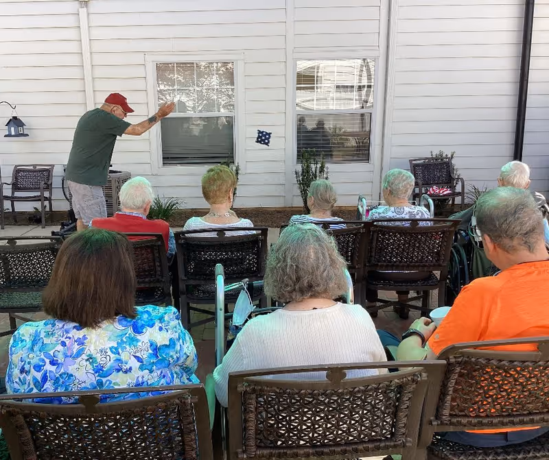 A group of elderly people seated outdoors on chairs facing a man who is standing and throwing a bean bag towards a target on the wall of a building. The setting appears to be a patio area with white siding on the building and some plants near the wall.
