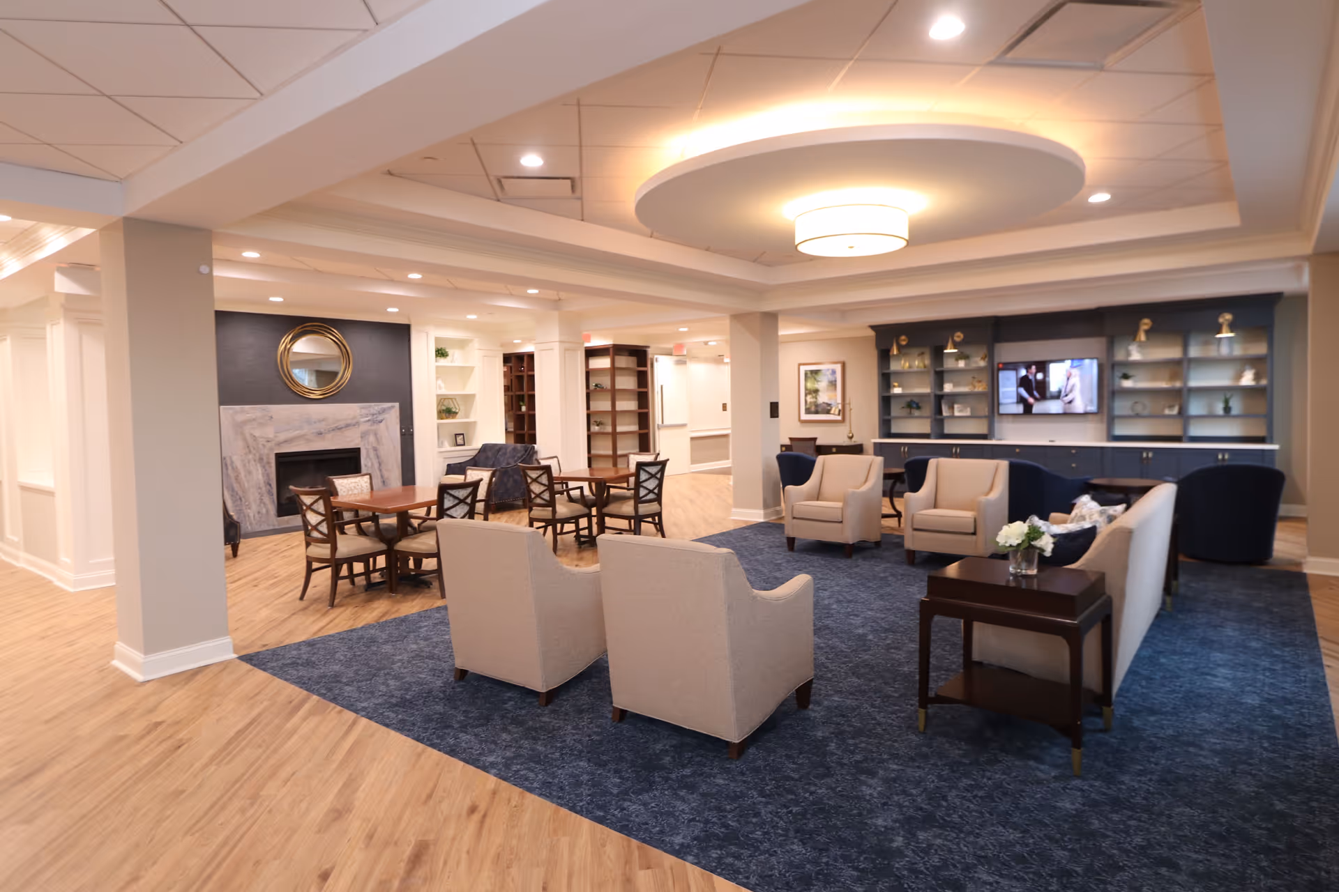 A spacious and well-lit senior living common area with beige armchairs and sofas arranged on a blue carpet. There are wooden tables and chairs near a fireplace with a round mirror above it. The room features built-in shelves and cabinets with decorative items and a TV mounted on the wall. The ceiling has recessed lighting and a modern circular light fixture.