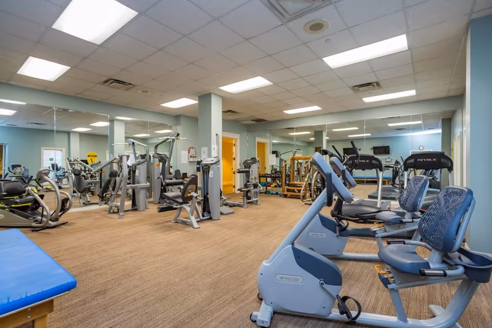 A spacious fitness room with various exercise equipment including stationary bikes, weight machines, and treadmills. The room has light blue walls, large mirrors covering one side, and a carpeted floor. Bright fluorescent lights illuminate the ceiling.