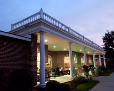 Exterior view of a brick building with a covered porch supported by white columns, illuminated at dusk with some people sitting on the porch and plants decorating the area.