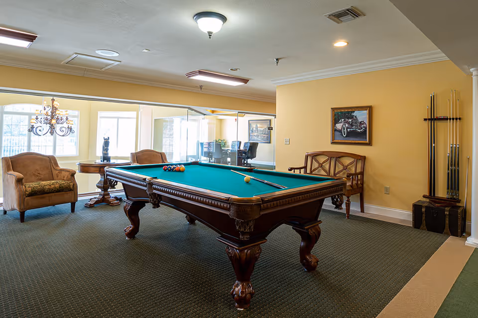 A well-lit interior room featuring a classic wooden pool table with billiard balls and a cue stick on it. The room has beige walls, carpeted floor, and several seating options including an armchair, a wooden bench, and a small round table with a decorative sculpture. A chandelier hangs near the windows, and a painting of a vintage car is mounted on the wall. Pool cues are stored in a rack in the corner.