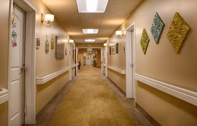 A well-lit hallway in an assisted living facility with beige walls, carpeted floor, wall-mounted handrails, decorative wall art, and multiple doors leading to rooms on either side.