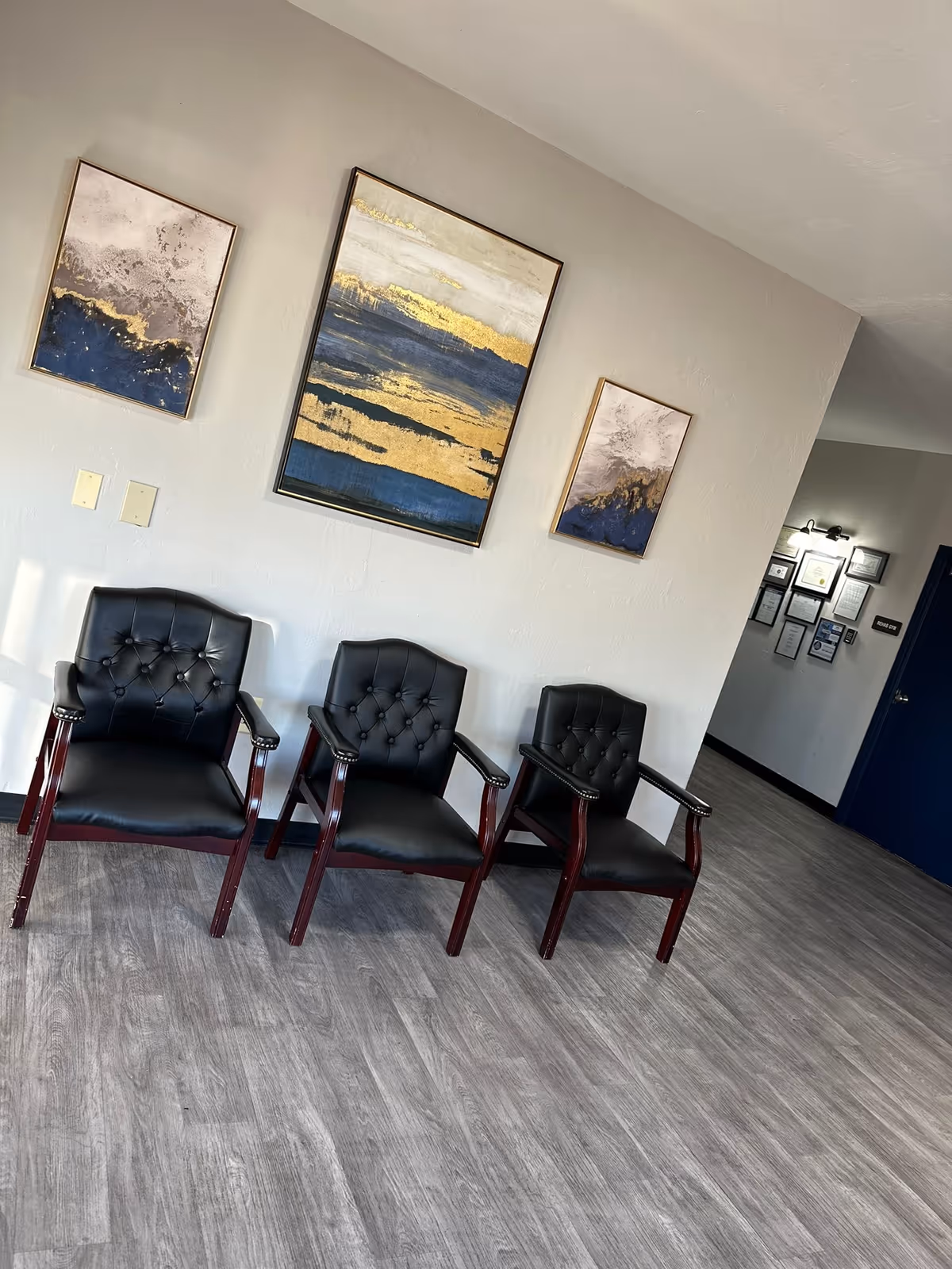 Three black leather chairs with wooden armrests and legs are arranged against a light gray wall. Above the chairs, there are three abstract paintings featuring blue, gold, and gray tones. The floor has a gray wood-like finish, and in the background, there is a hallway with framed certificates or documents on the wall and a dark blue door.