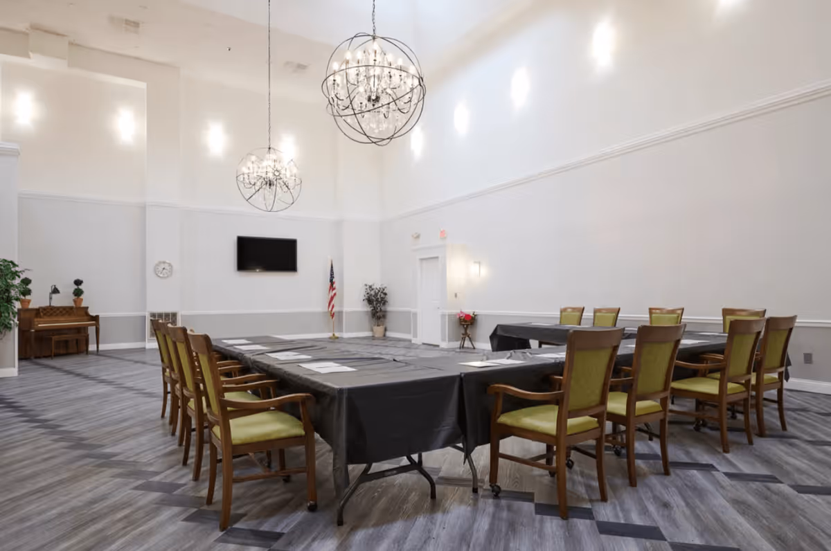 A large, bright meeting room with high ceilings and two spherical chandeliers. The room has a U-shaped arrangement of tables covered with black tablecloths and surrounded by wooden chairs with green cushions. There is a piano and some plants in the corner, a wall-mounted TV, an American flag, and a clock on the wall.