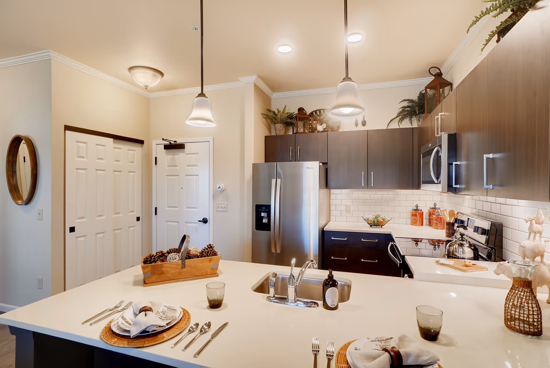 Modern kitchen interior with a large white island countertop set for two with plates, cutlery, and glasses. The kitchen features dark wood cabinets, a stainless steel refrigerator, microwave, and stove. Two pendant lights hang above the island. Decorative plants and kitchen items are placed on top of the cabinets and counters.