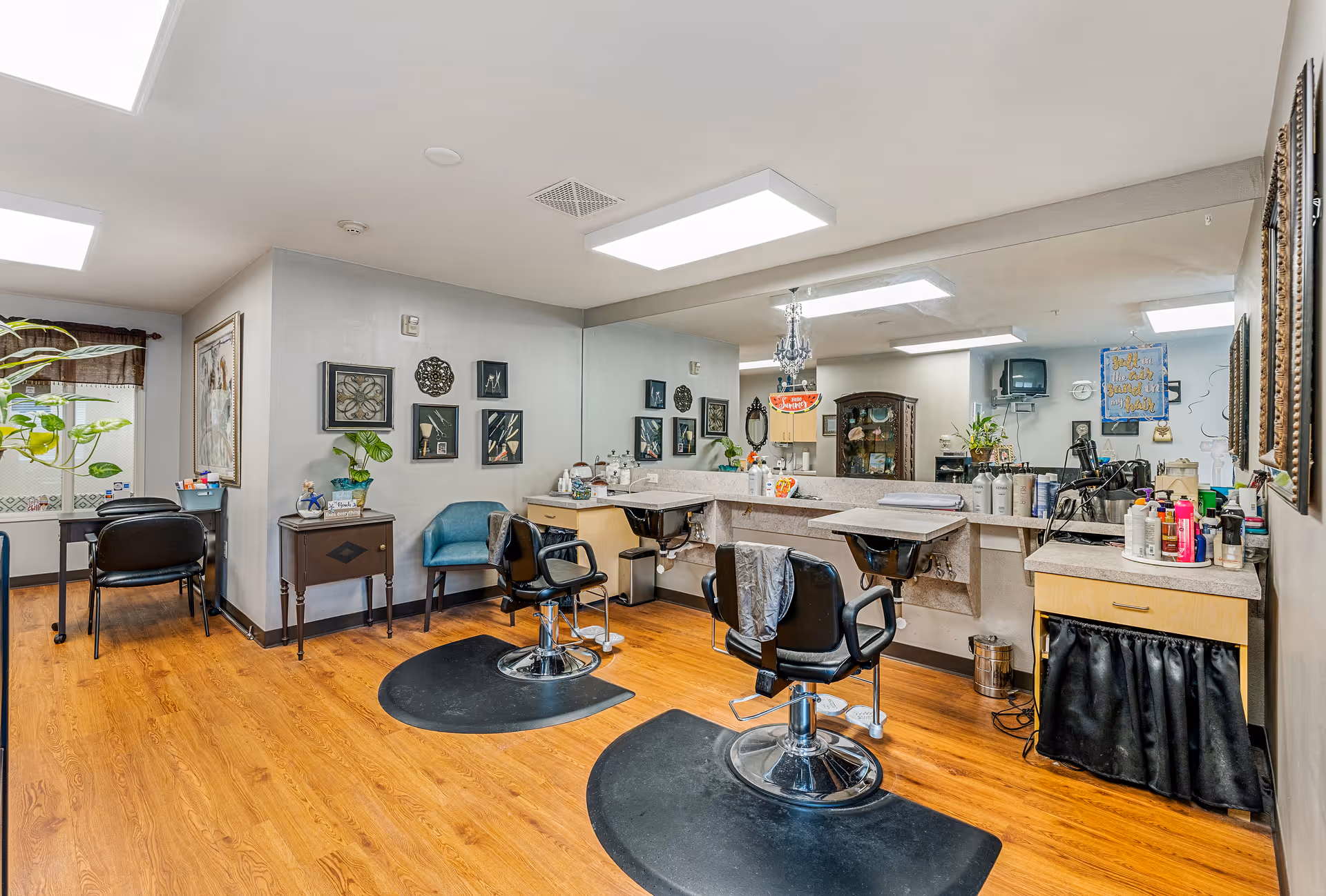 Interior view of a hair salon area with two black salon chairs on black mats in front of a large mirror and countertop with various hair care products. The room has wooden flooring, light gray walls decorated with framed artwork, and a small seating area with a blue chair and a small table. There is a window with a valance and a plant near the seating area.