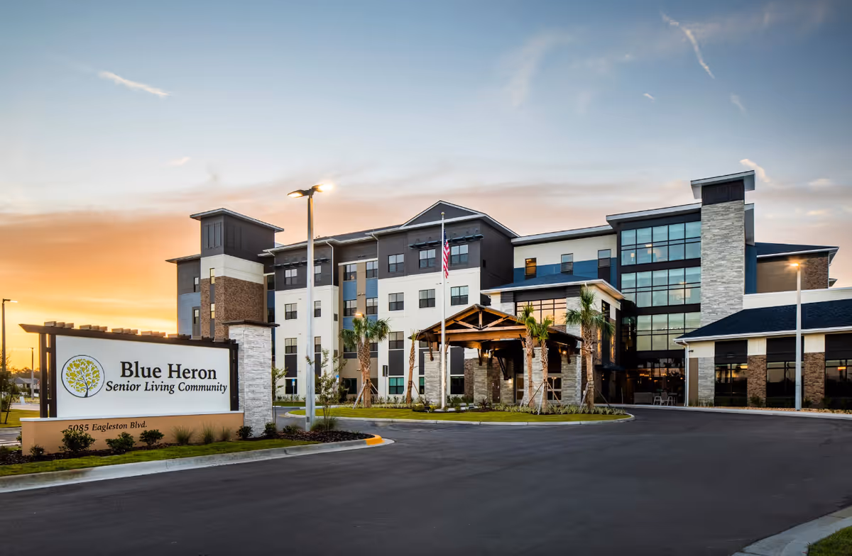 Exterior view of Blue Heron Senior Living Community building at sunset with a large sign in front displaying the facility name and address. The building is modern with multiple floors, large windows, palm trees, and a covered entrance.