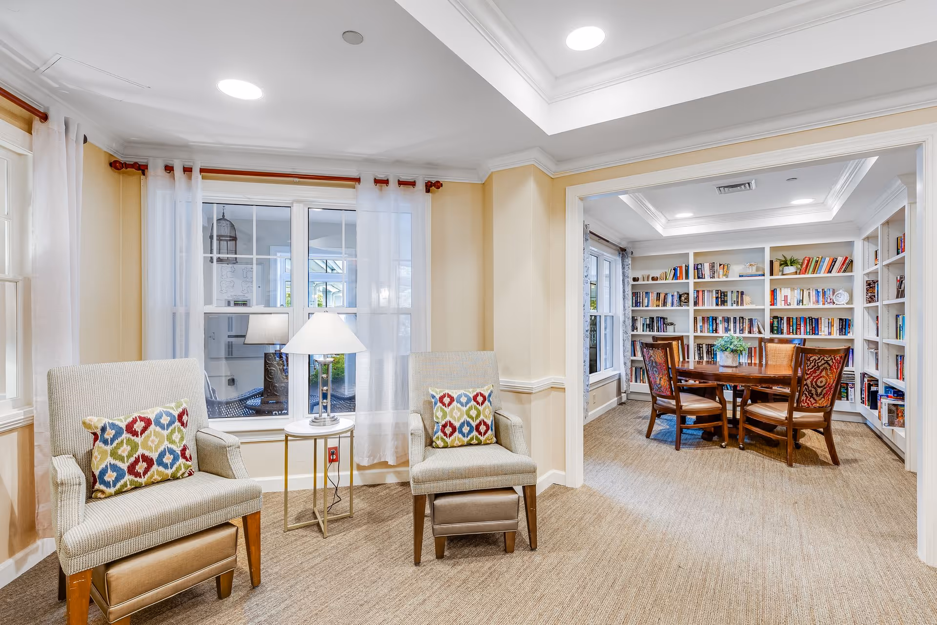 A cozy interior room with two cushioned armchairs featuring colorful patterned pillows and a small round side table with a lamp between them. In the background, there is a separate room with a wooden round table surrounded by four chairs, and white built-in bookshelves filled with books and decorative items. The walls are painted a soft beige, and the room is well-lit with recessed ceiling lights and natural light coming through the windows with sheer white curtains.