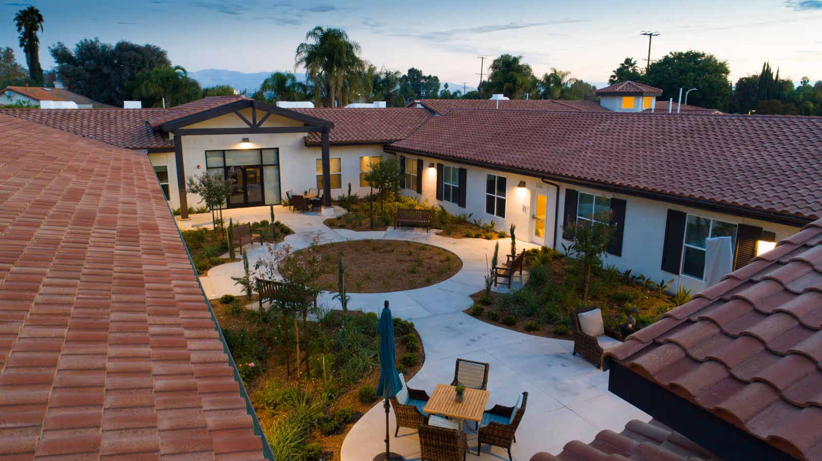 Outdoor courtyard area of The Preserve at Woodland Hills Assisted Living & Memory Care with a circular garden in the center, surrounded by paved walkways, benches, chairs, tables, and plants. The building has a red tiled roof and white walls with windows and doors opening to the courtyard. Trees and mountains are visible in the background during dusk.