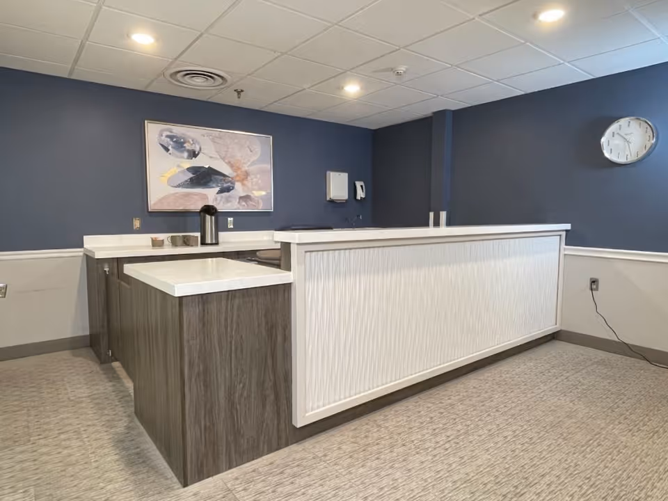 Reception desk area with a modern design featuring a white textured front panel and wood finish sides. The wall behind the desk is painted dark blue and has a framed abstract artwork. A clock is mounted on the right wall, and the ceiling has recessed lighting.