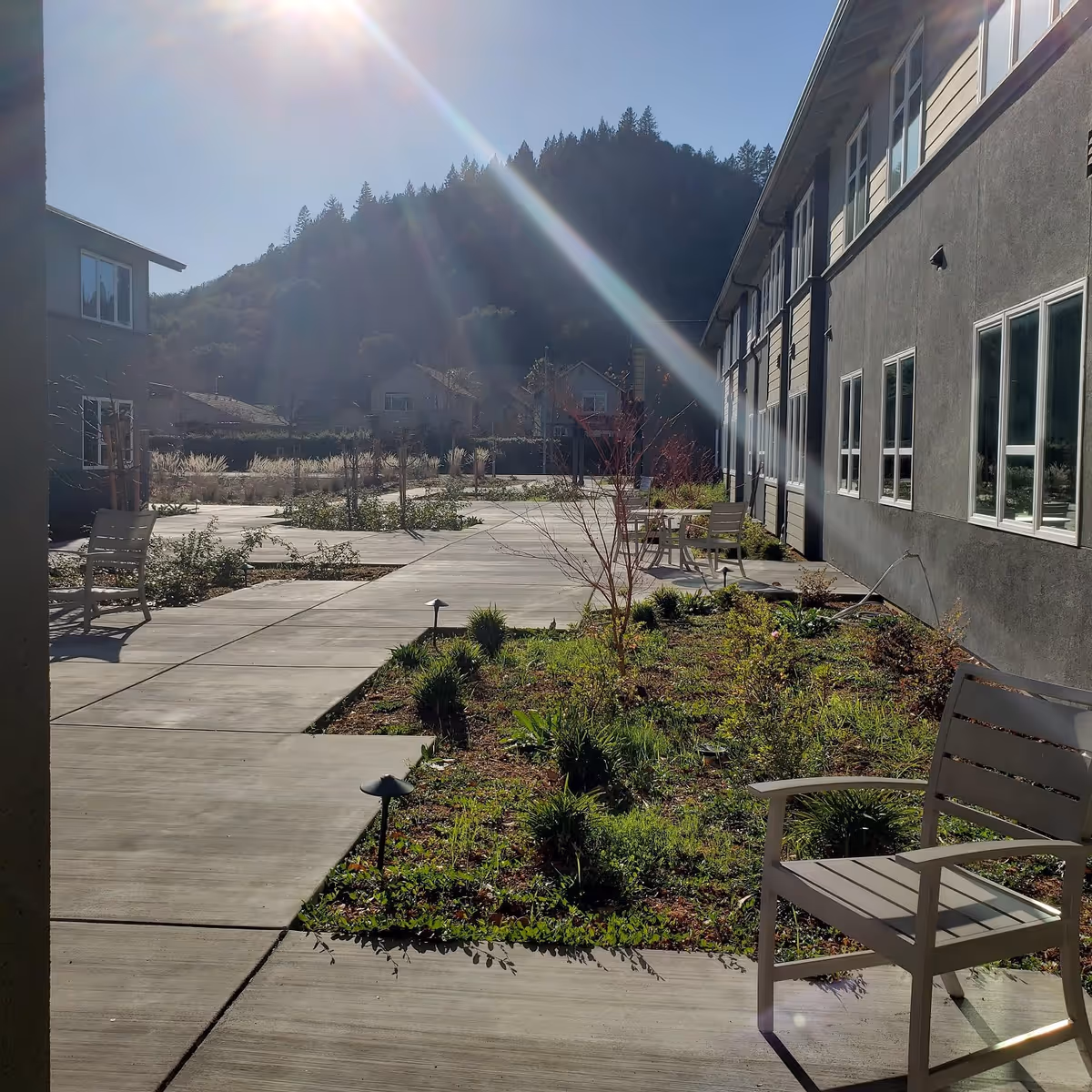 Outdoor courtyard area of a senior living facility with concrete walkways, planted garden beds, several chairs, and a backdrop of forested hills under a clear blue sky with sunlight streaming in.