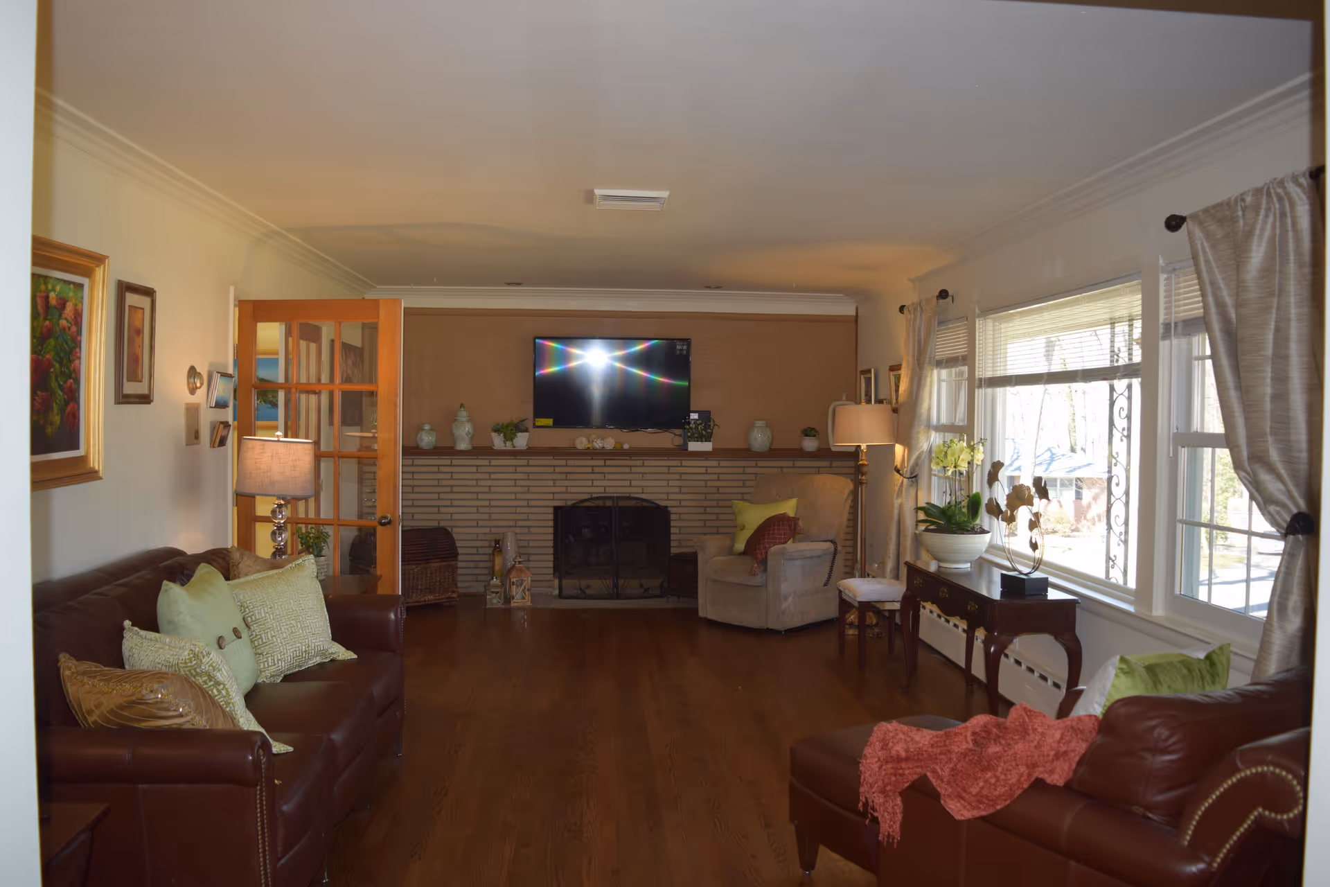 A cozy living room with wooden flooring, two brown leather sofas adorned with green and beige cushions, a beige armchair with pillows, a wooden side table with a lamp, and a large window with curtains letting in natural light. A flat-screen TV is mounted above a brick fireplace decorated with vases and plants.